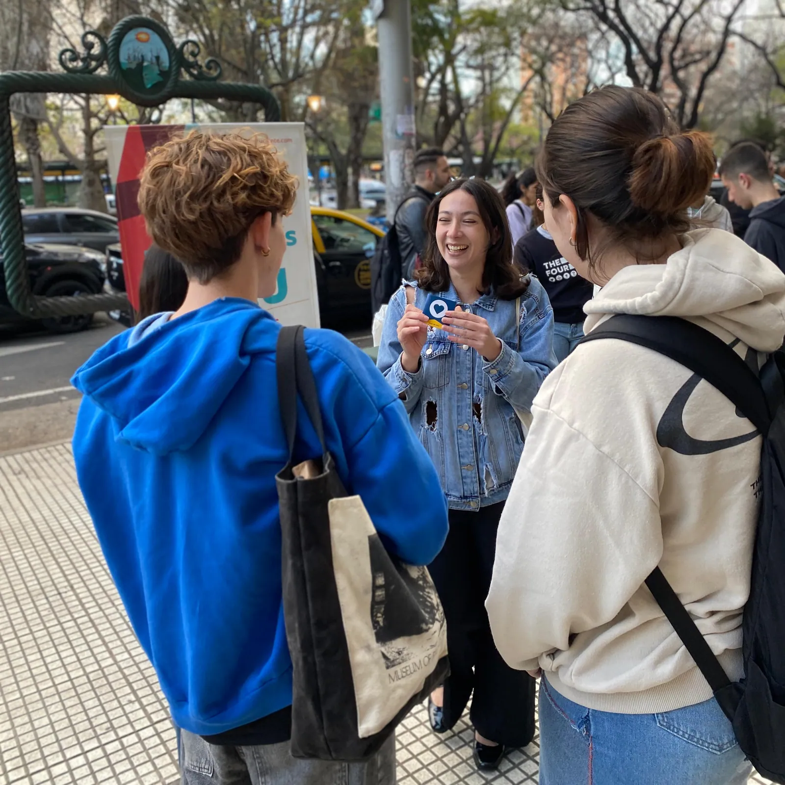 Tres estudiantes conversando en la vereda; una de ellas sostiene un folleto y sonríe